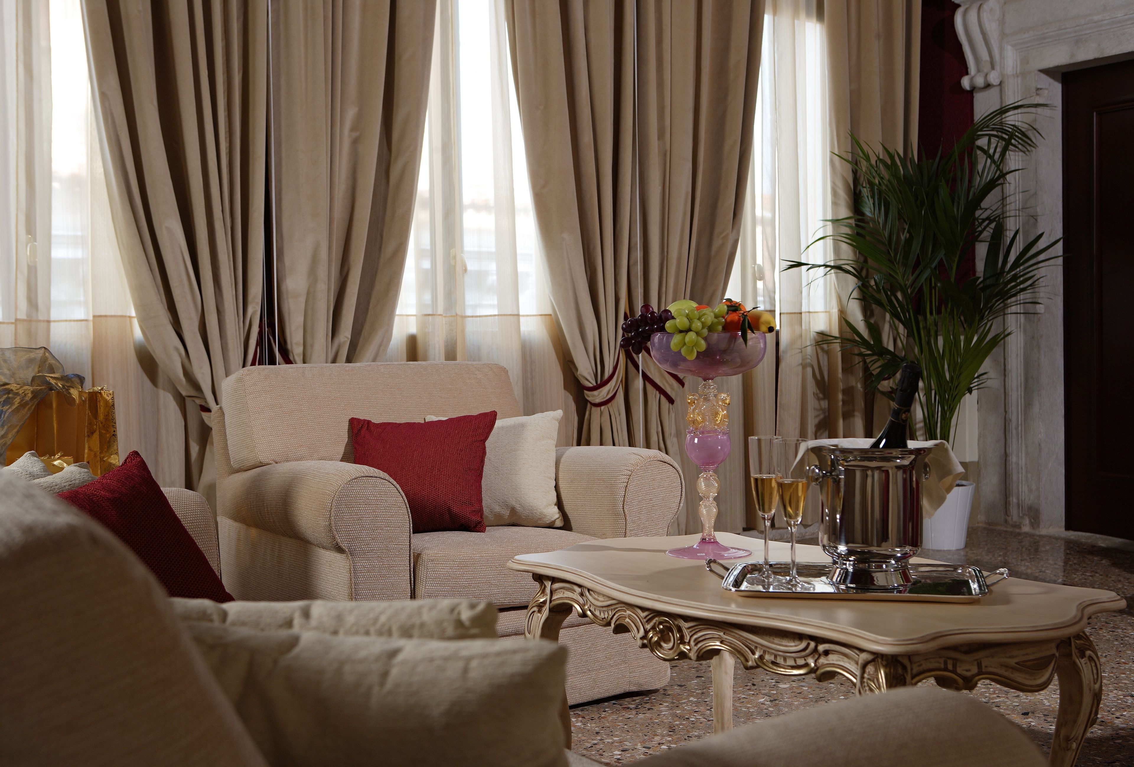 A living room with a beige sofa, red cushions, a coffee table with a silver pitcher and glasses, and a vase with fruit.