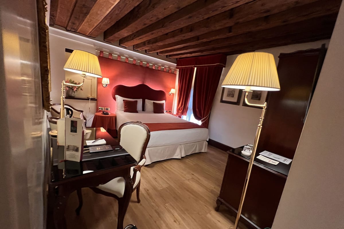 A hotel room with a wooden ceiling, a bed with red curtains, a desk with a lamp, and a chair.