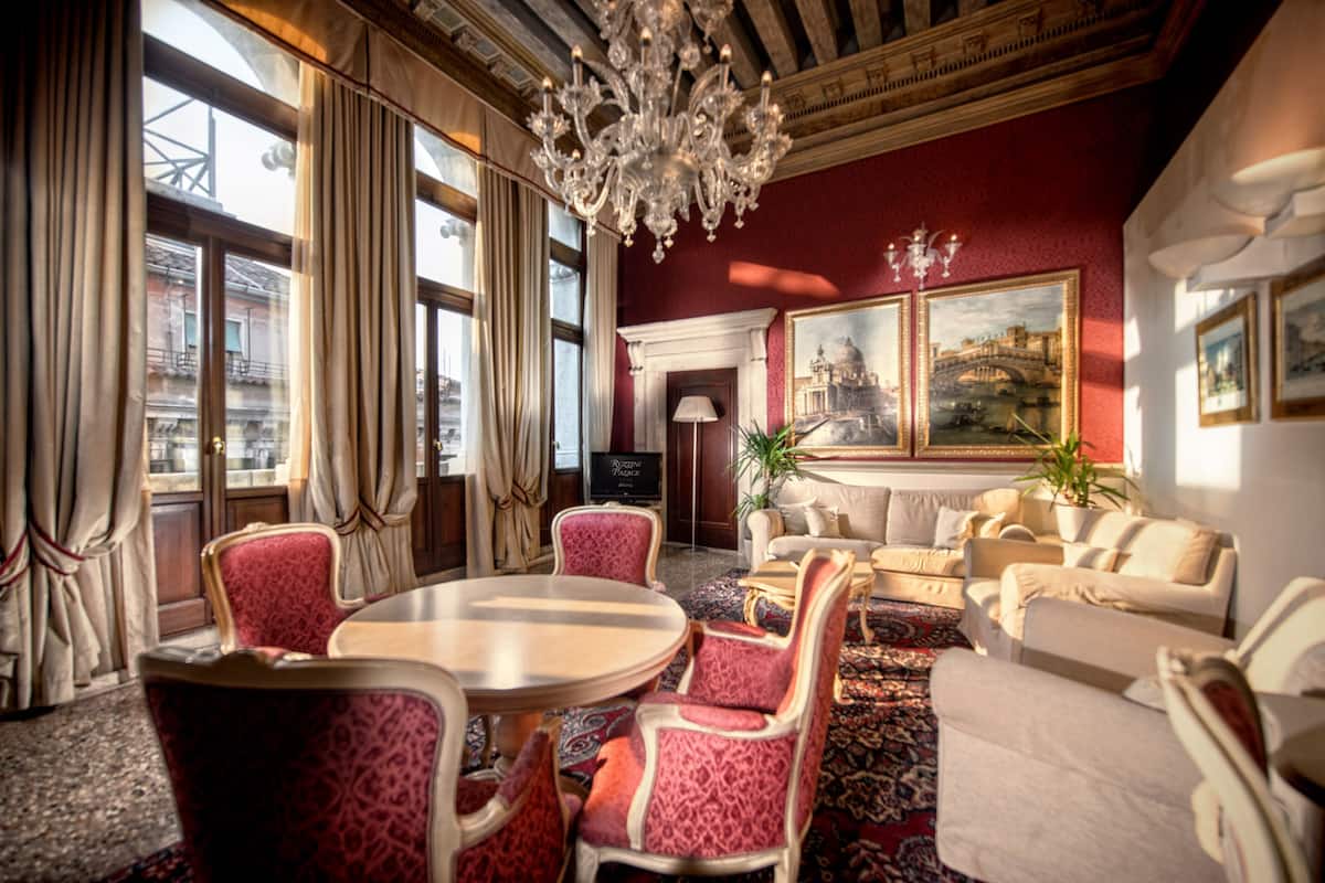 A hotel lobby with a round table, red upholstered chairs, a sofa set, a chandelier, and large windows with curtains.
