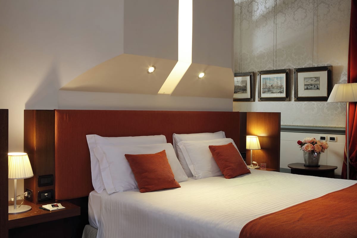 A hotel room with a large bed, two bedside tables, a nightstand, a lamp, a vase with flowers, and framed pictures on the wall.