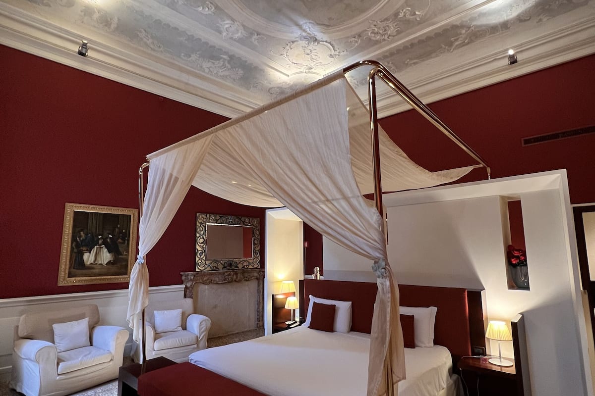 A luxurious bedroom with a canopy bed, a red accent wall, and ornate ceiling details.