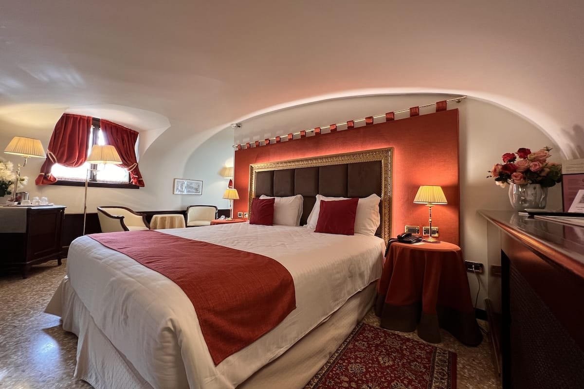A hotel room with a large bed, a desk, a chair, a nightstand, and a vase with flowers.