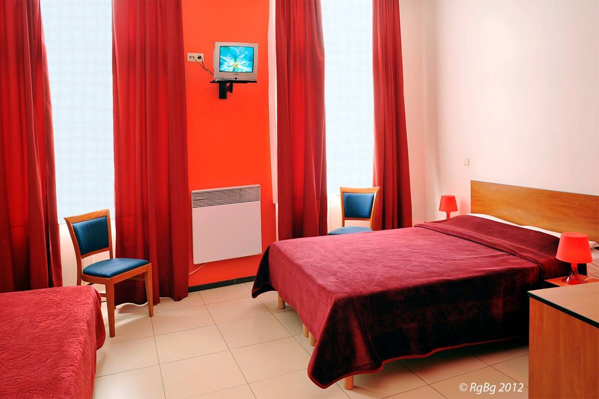 Una camera d'albergo con un letto, due poltrone, una scrivania, un televisore e tende rosse.