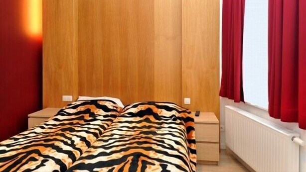 A bedroom with a wooden headboard, a bed with a zebra-patterned blanket, two bedside tables, and red curtains. - Hotel The Moon (Brussels)