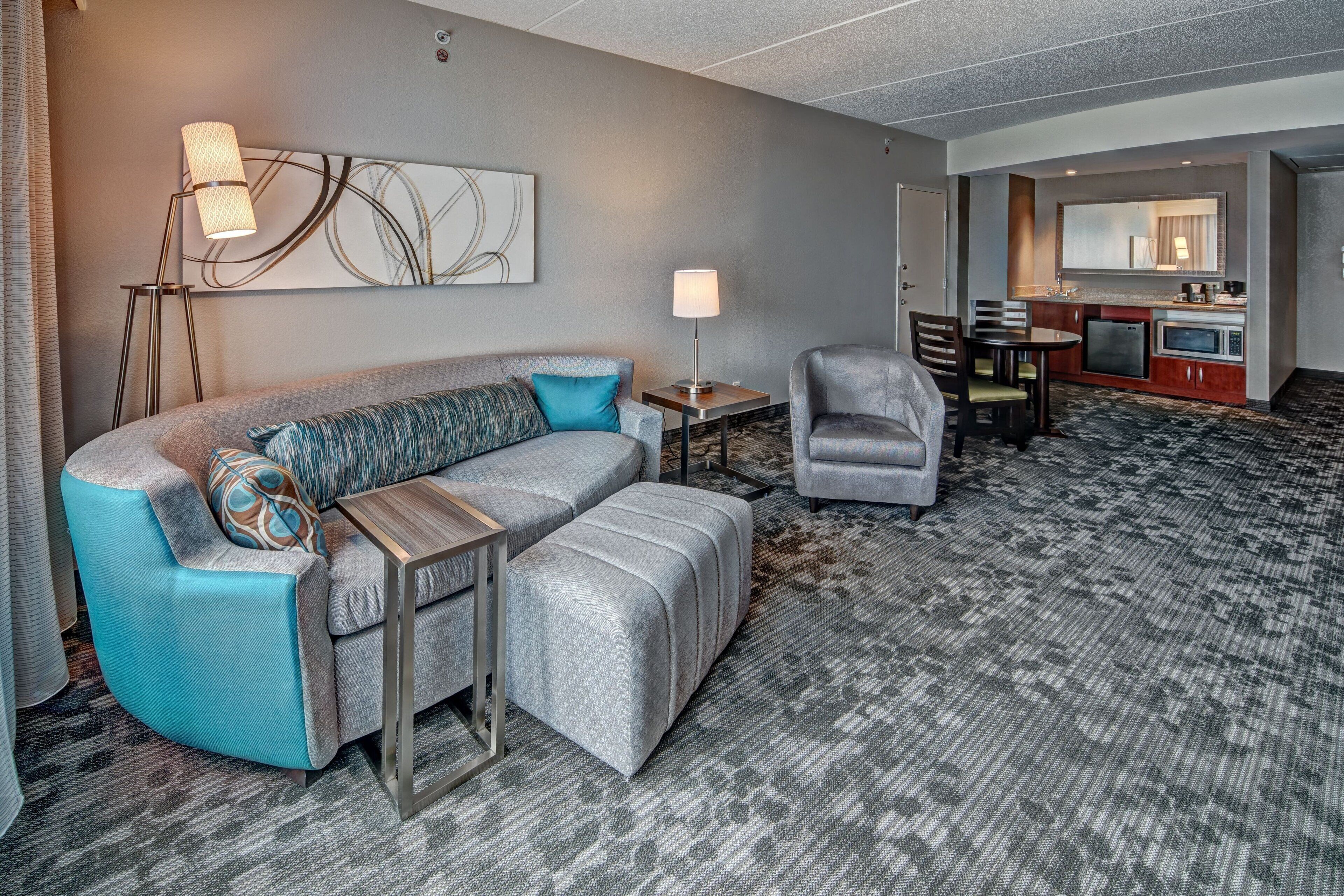 A modern hotel room with a grey sofa, a grey armchair, a small table, a lamp, and a kitchenette.