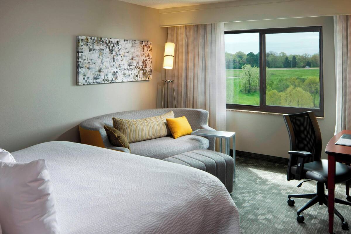 A hotel room with two beds, a desk, a chair, a lamp, and a view of a green landscape.