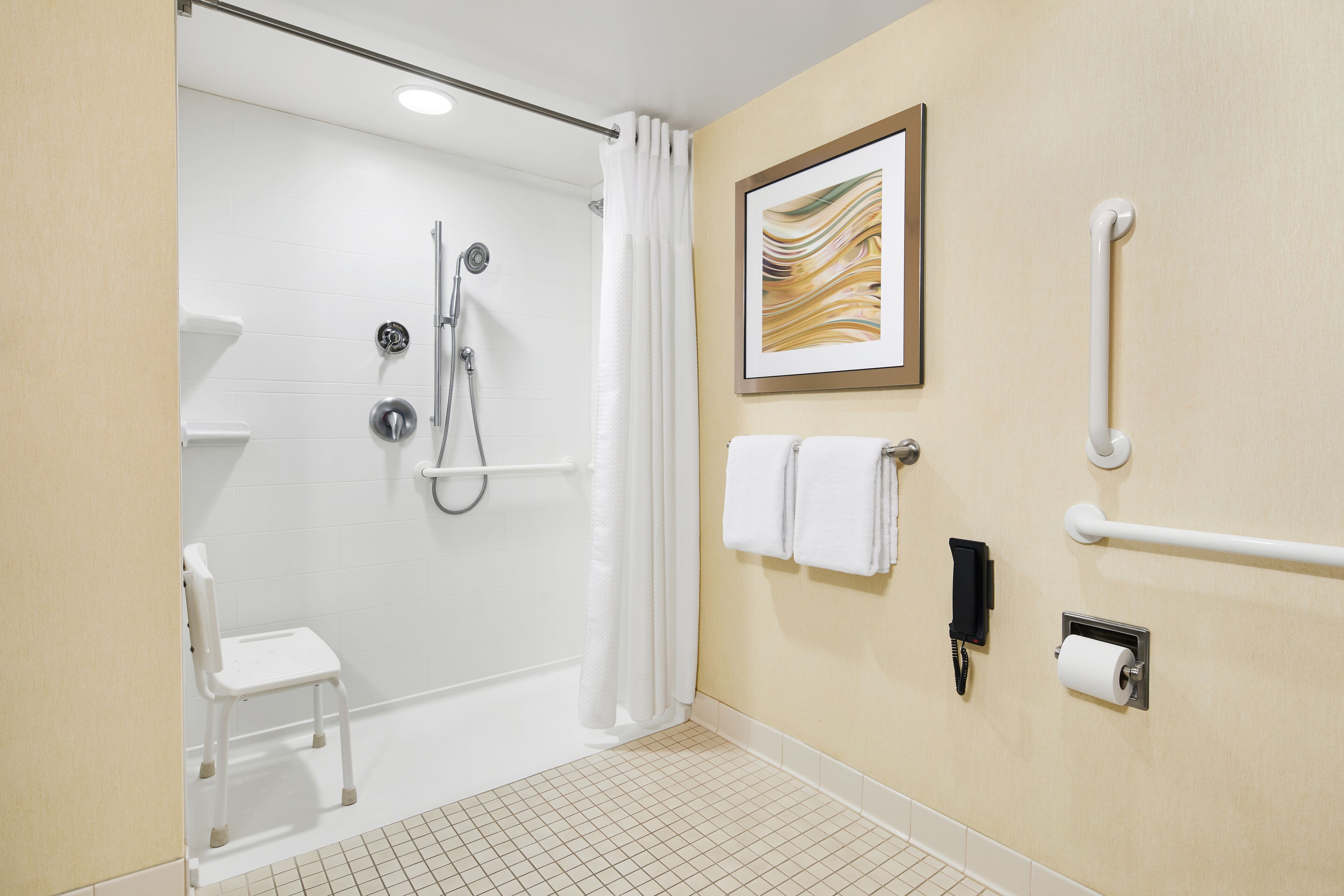 A bathroom with a shower, a toilet, a chair, a towel rack, a phone, and a picture on the wall.