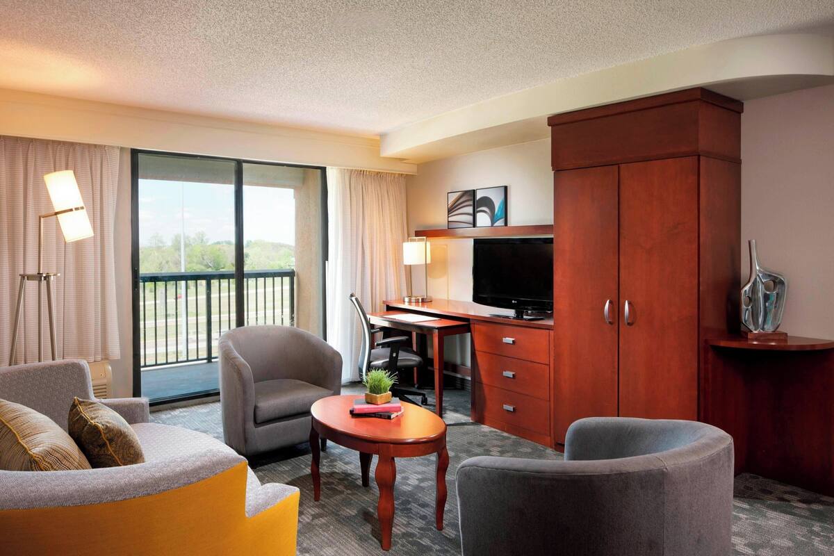A hotel room with a large window, a desk, a TV, and a seating area with two chairs and a sofa.