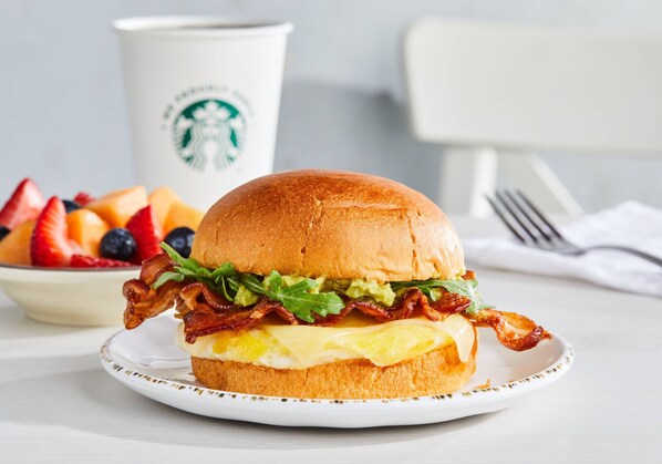 Un sándwich de desayuno sobre un plato, con una taza y un bol de fruta al fondo.
