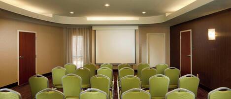 Un salón de conferencias con filas de sillas verdes, una pantalla de proyección grande y una puerta de madera a la derecha.