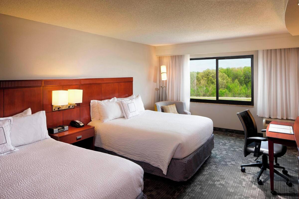 A hotel room with two beds, a desk, a chair, and a large window with a view of greenery.