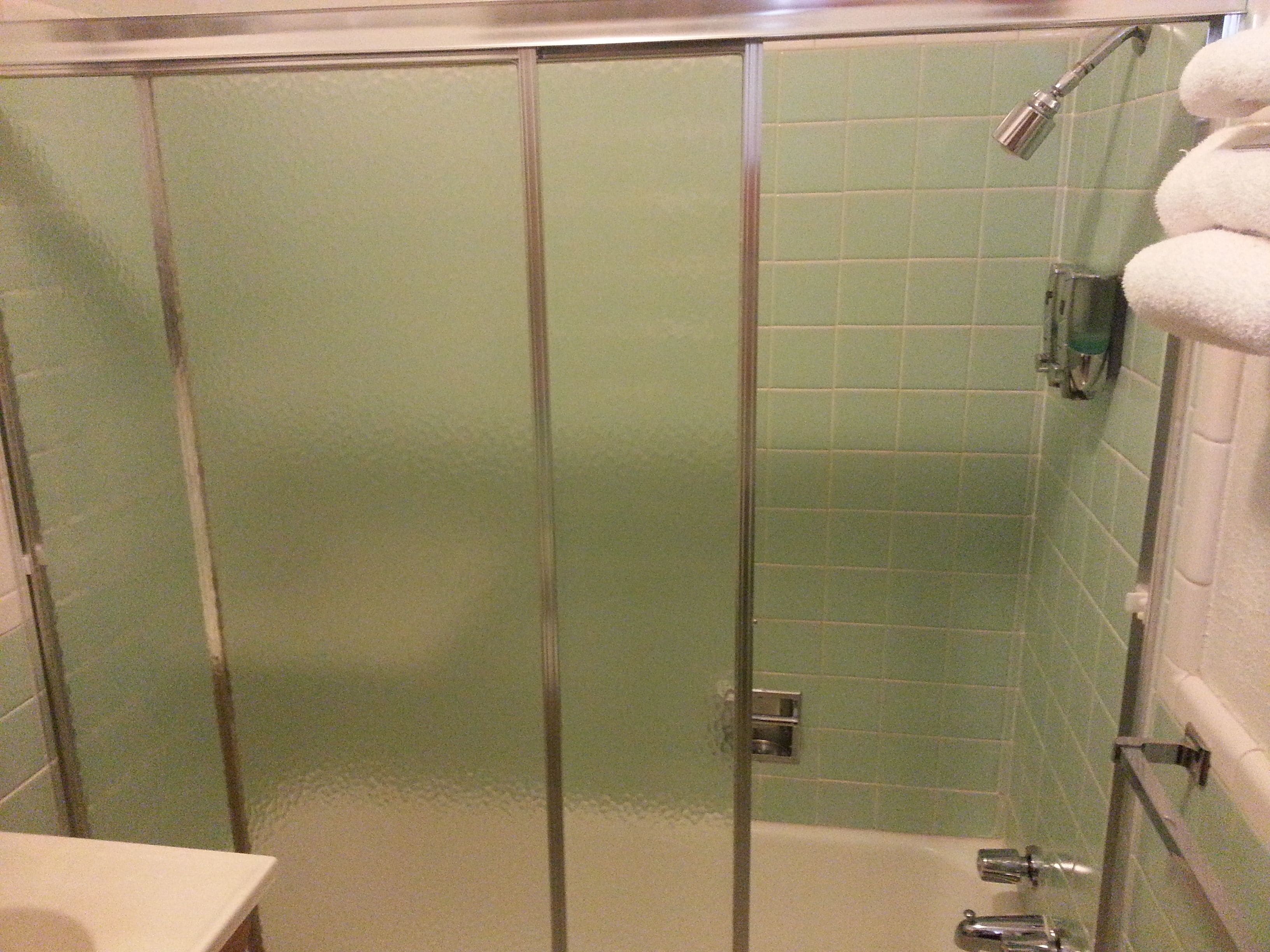 A glass-enclosed shower stall with a green tiled wall and a white bathtub with a towel rack.