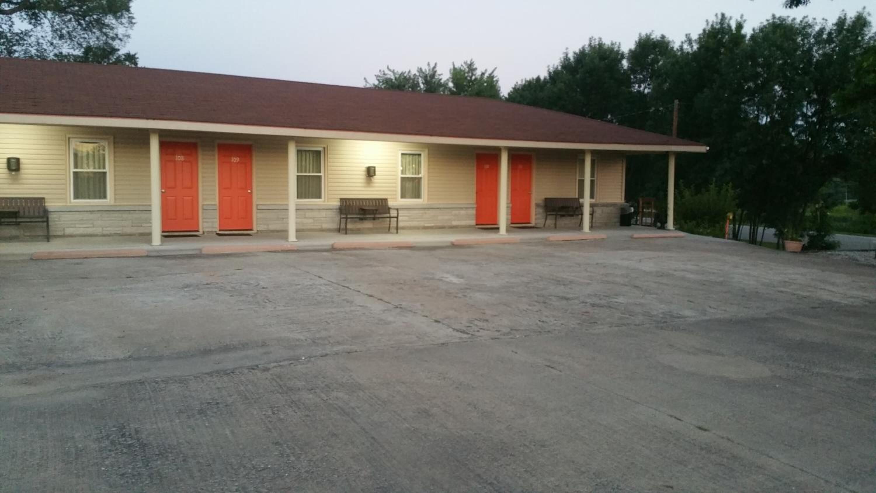 A motel with a red roof, beige exterior, and multiple red doors. There are benches outside and a paved parking area.