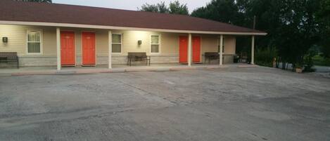 A motel with a red roof, beige exterior, and multiple red doors. There are benches outside and a paved parking area.