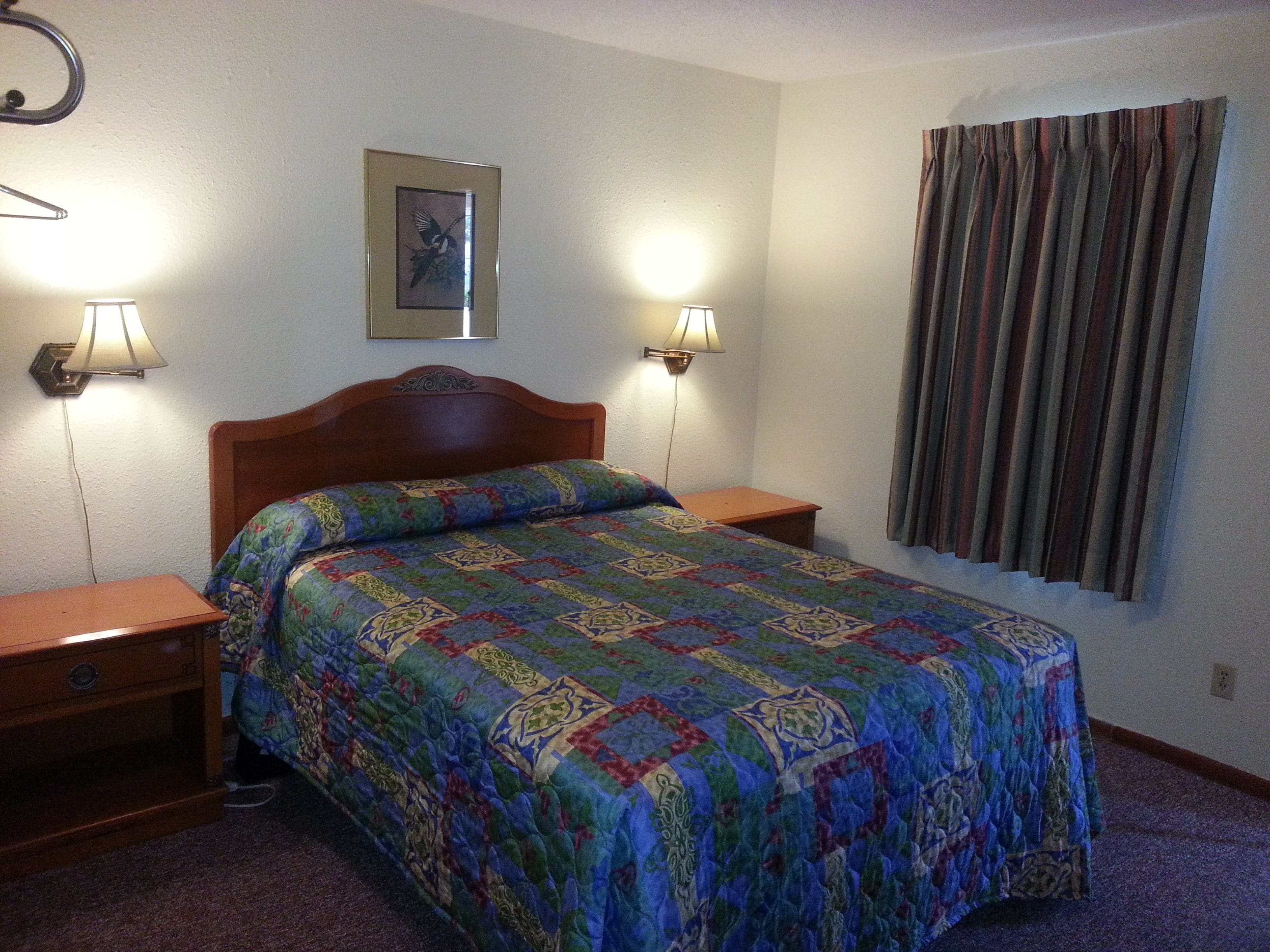 A neatly made bed with a colorful quilt, two bedside lamps, a framed picture on the wall, and a window with curtains.