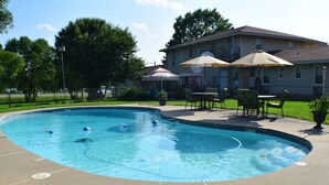 A swimming pool with a curved edge, surrounded by a concrete deck. There are outdoor chairs and umbrellas, and a house is visible in the background.