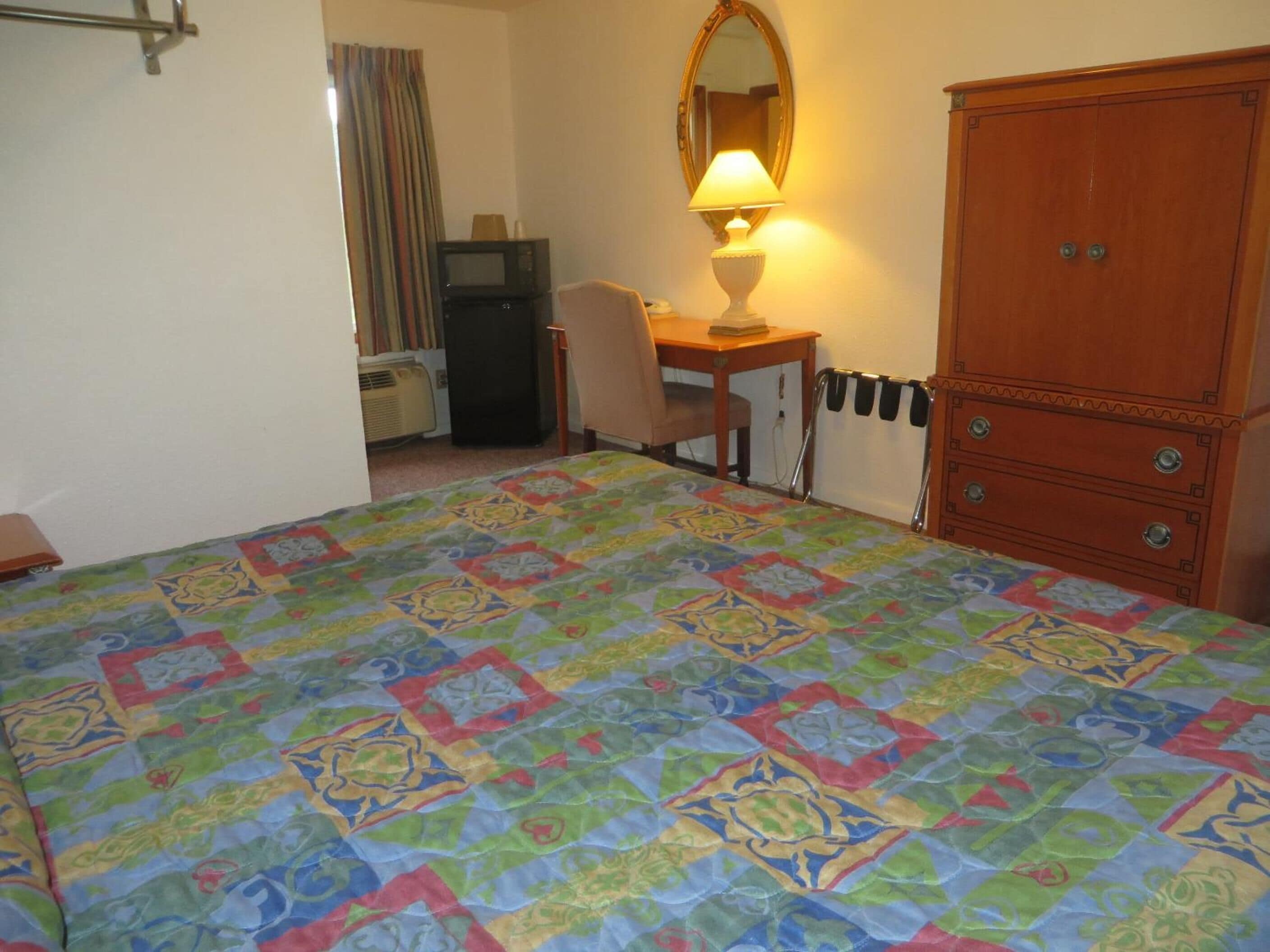 A hotel room with a bed, a desk, a chair, a wardrobe, a mirror, and a lamp.