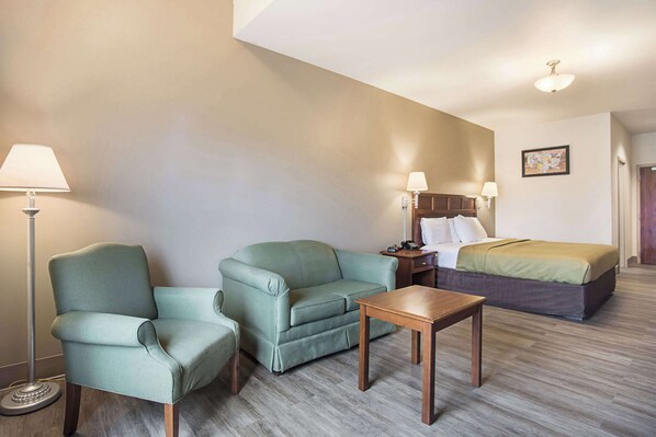 Una habitación de hotel con un sofá verde, una mesa de madera, una cama con cubrecama verde y marrón, y dos lámparas fijadas a la pared.