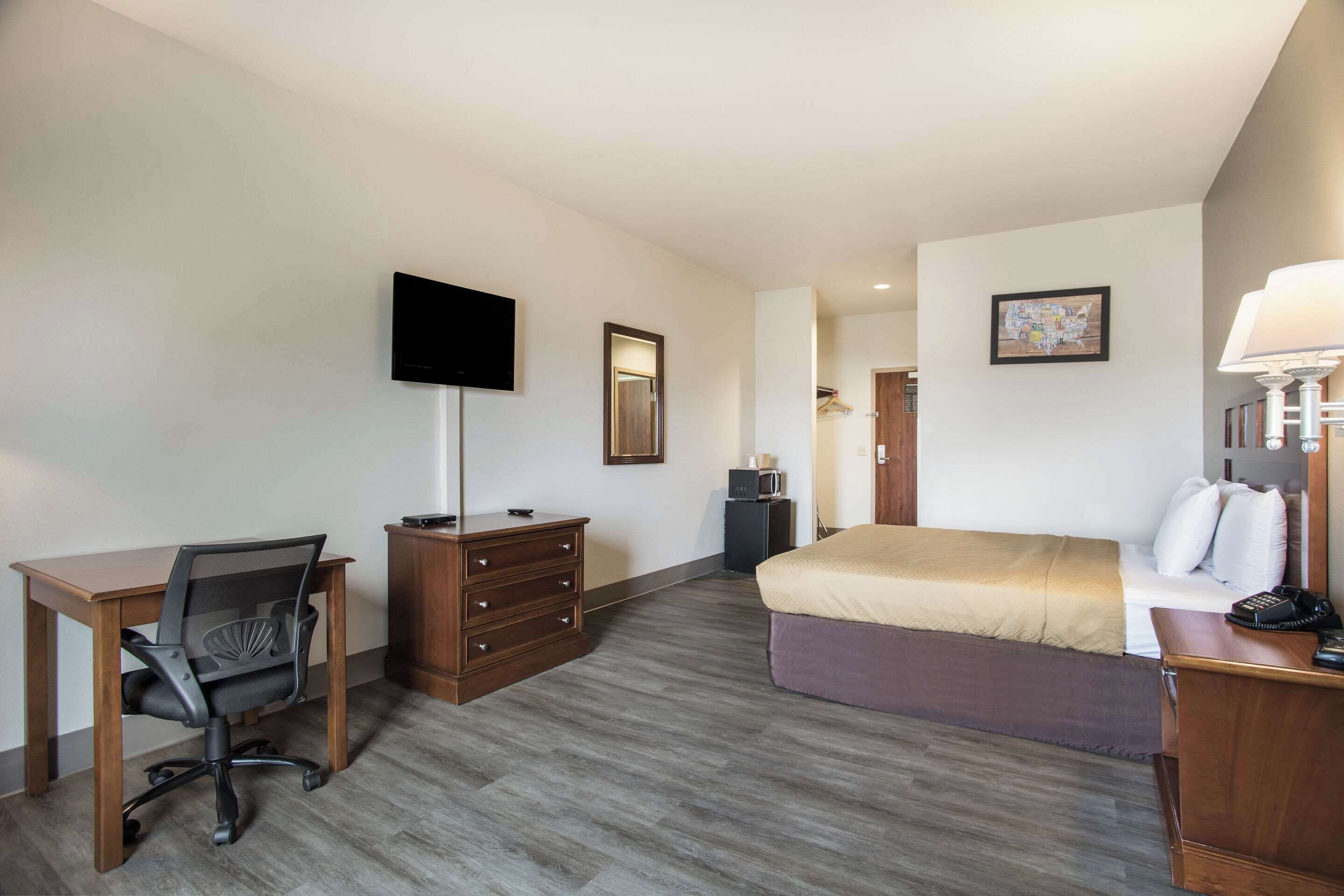 Quarto de hotel com cama, escrivaninha, cadeira, cômoda, televisão e abajur.