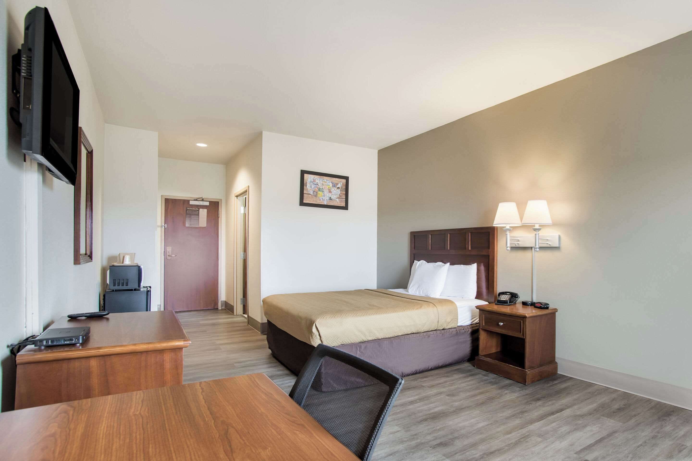 A hotel room with a bed, desk, chair, nightstand, lamp, and a television mounted on the wall.