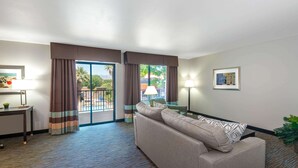 A hotel room with a sofa, a desk with a lamp, a large window with curtains, and a view of the outdoors. - Wingate by Wyndham St. George (St. George)