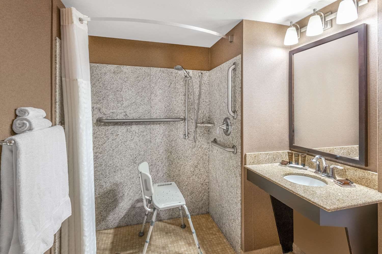 Une salle de bain équipée d’une douche, de barres d’appui, d’un banc accessible aux personnes en fauteuil roulant, d’un lavabo avec des articles de toilette et d’un grand miroir.