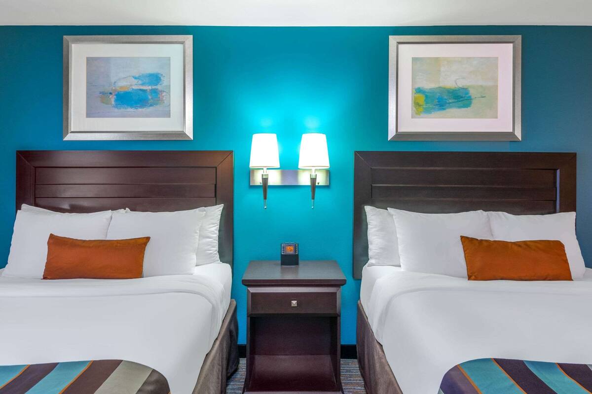 A hotel room with two beds, a nightstand with two wall-mounted lamps, and artwork on the walls.