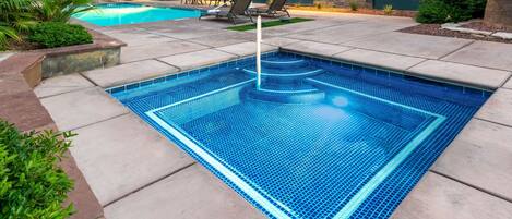 A rectangular pool with blue mosaic tiles, surrounded by a concrete deck with lounge chairs and a palm tree.