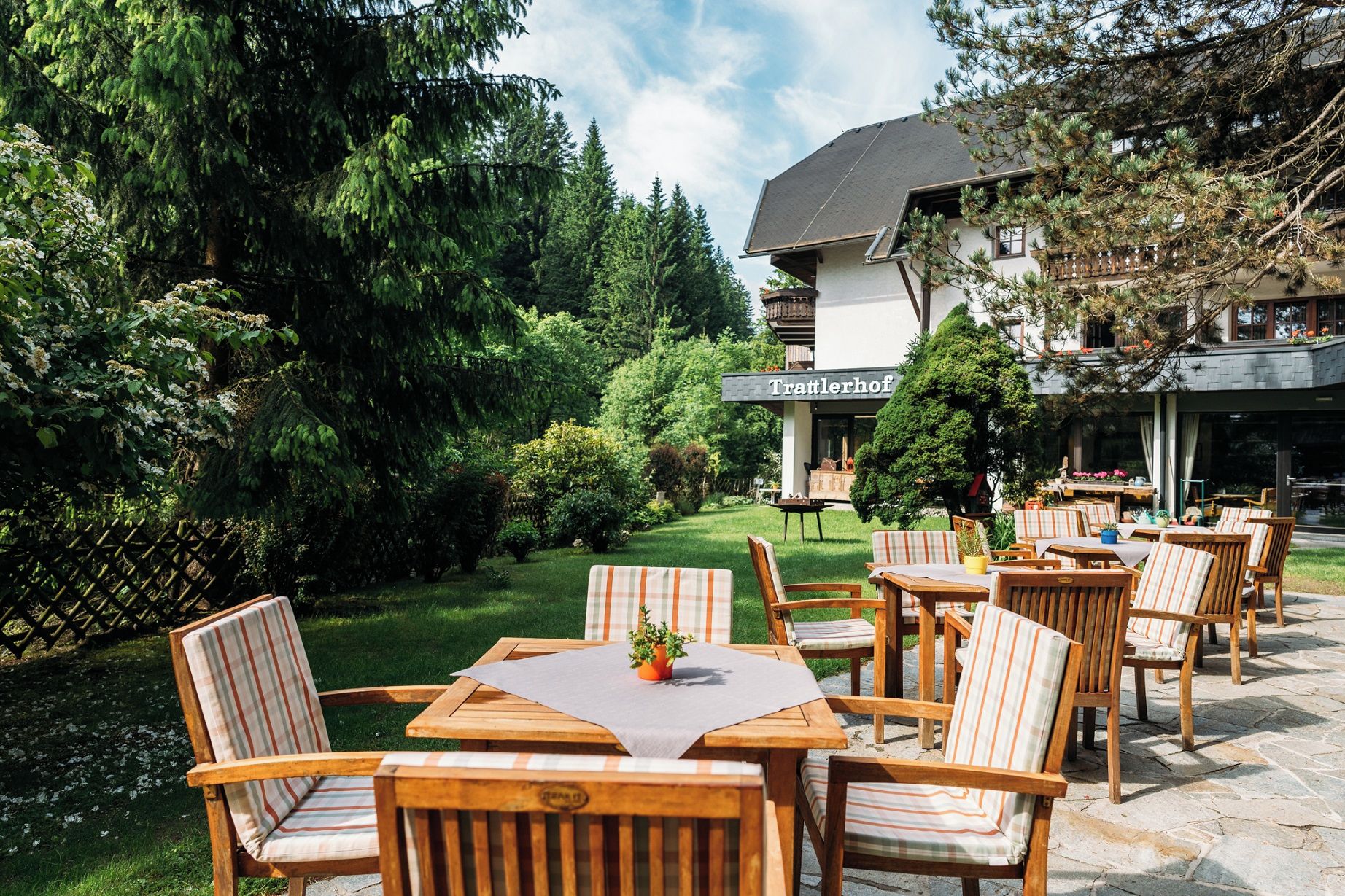 An outdoor seating area with wooden tables and chairs, a building with a sign reading 'Trattlerhof,' and a lush garden with trees and shrubs.