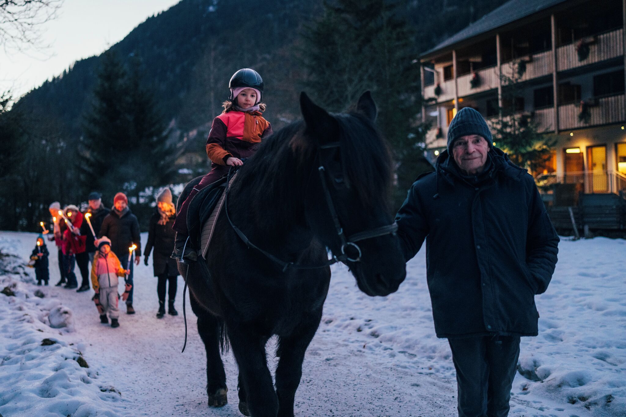 A horse and rider walking on a snow-covered path with a building in the background.