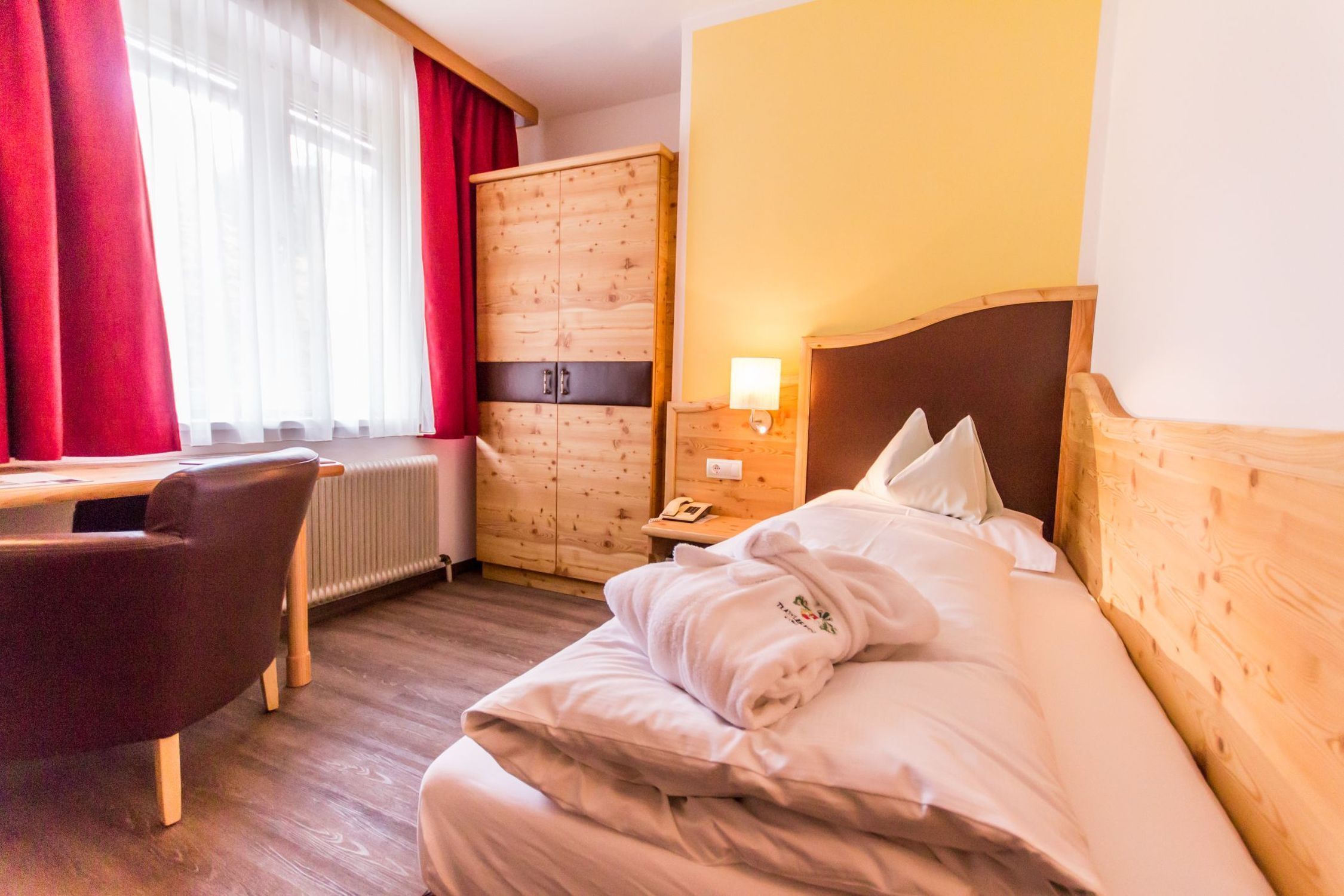 Una habitación de hotel con cama, escritorio, silla, armario y mesita de noche con lámpara.