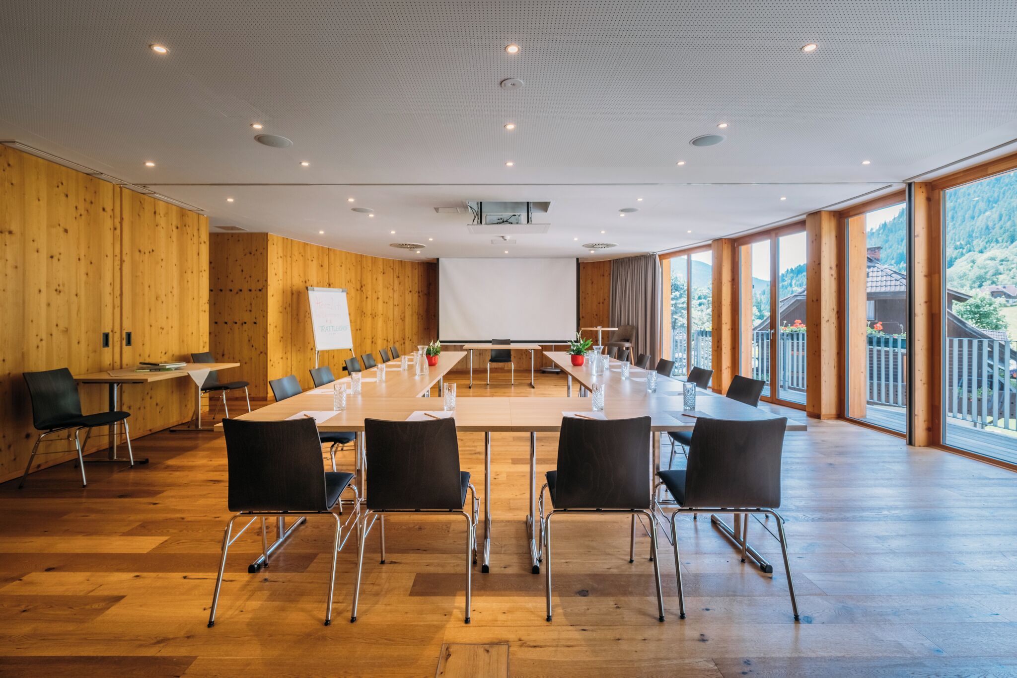 Un salón de conferencias con una larga mesa, sillas negras, paredes de madera y ventanas amplias.