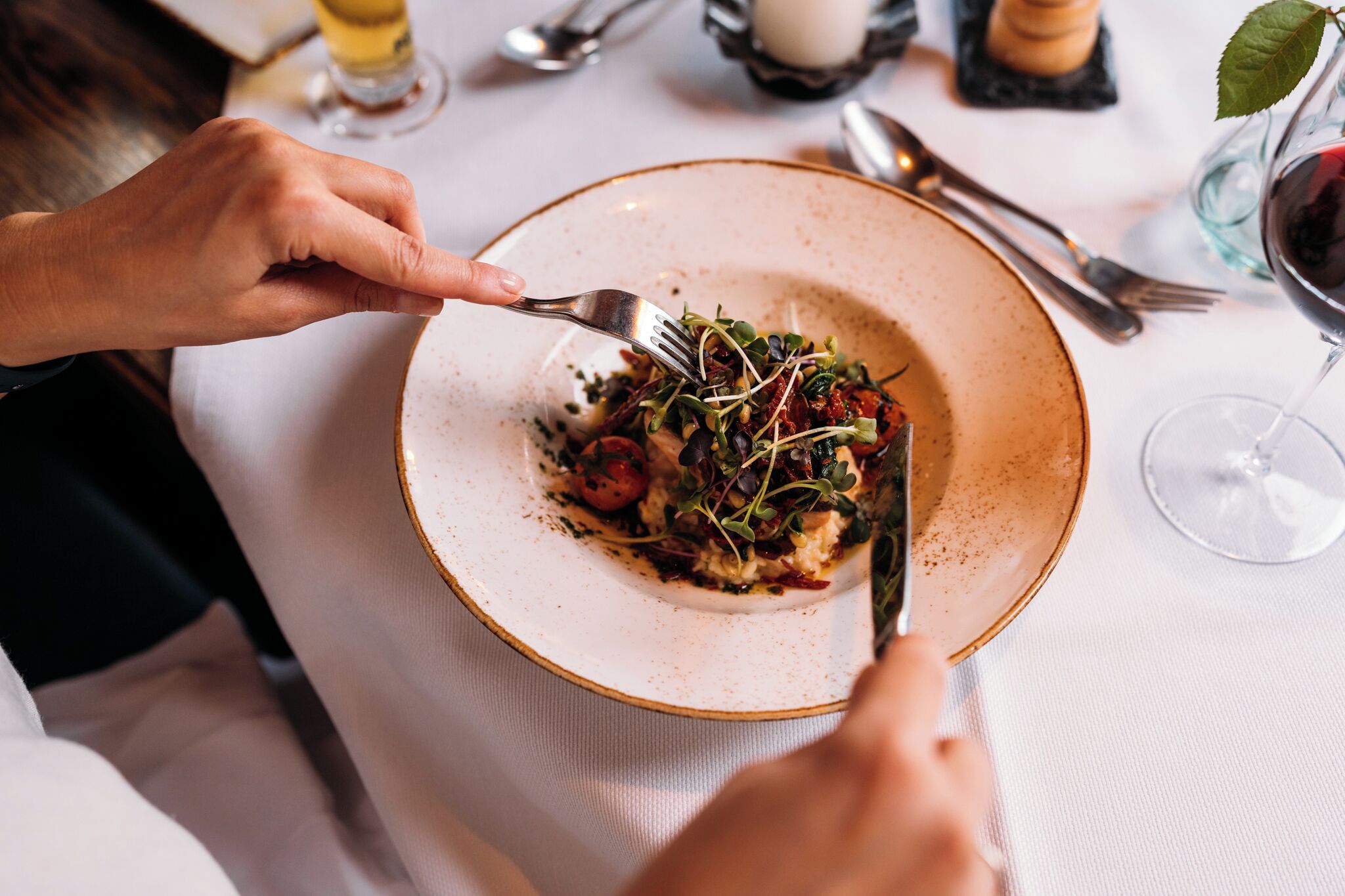 A person dining at a table with a plate of food, cutlery, and a glass of red wine.