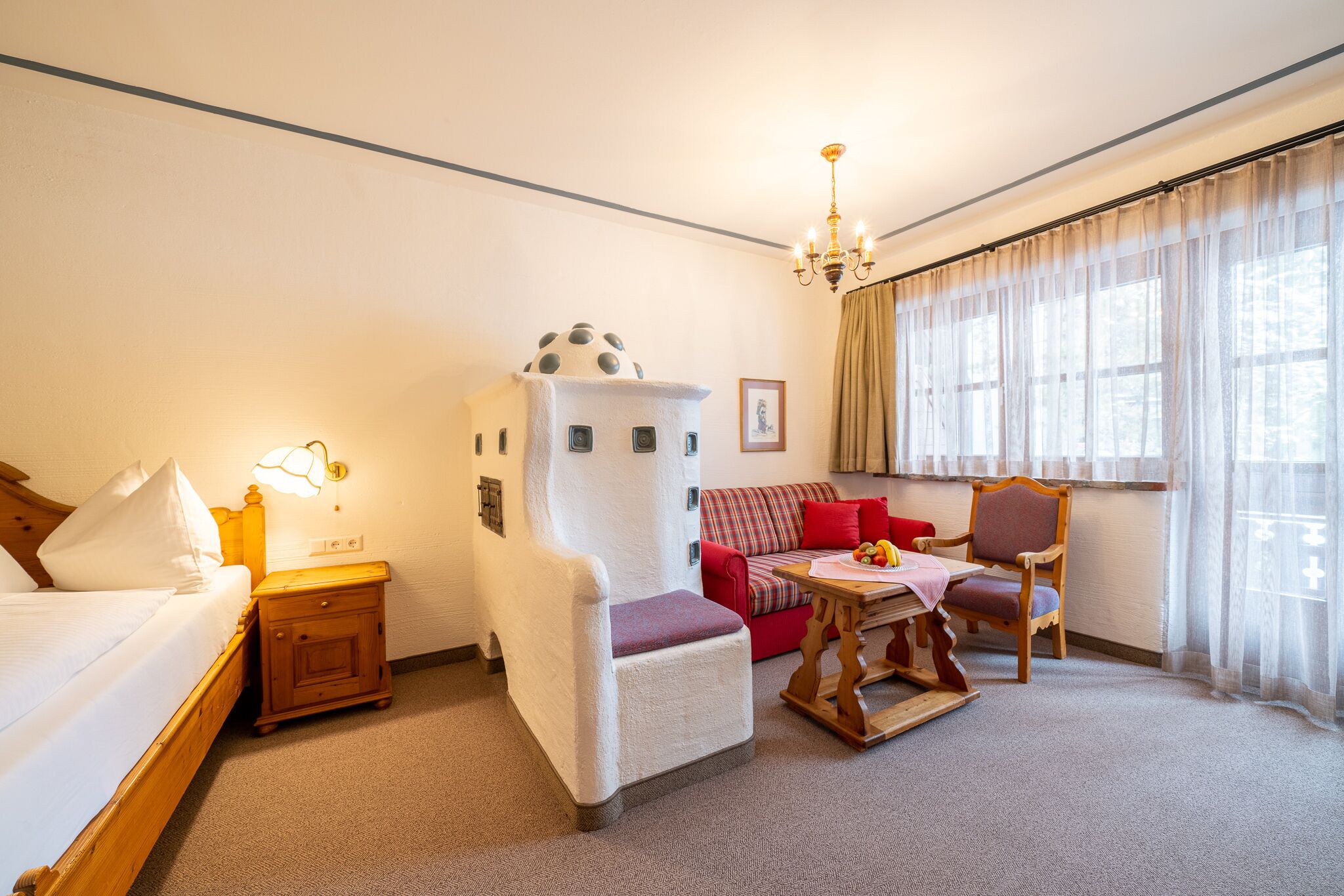 Una habitación de hotel con cama, un sillón blanco, un sofá rojo, una mesa de madera y una araña.