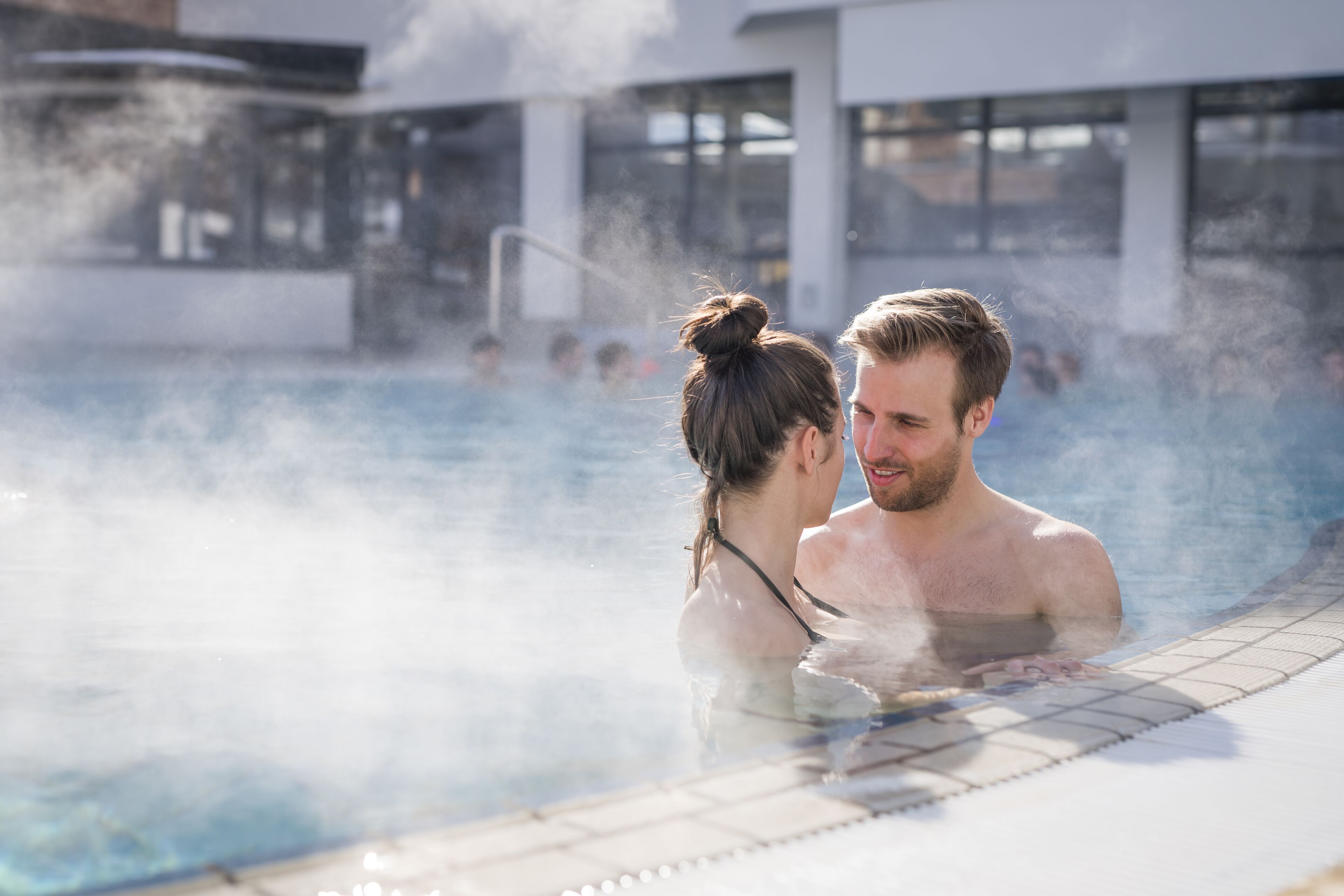 A couple in a hot tub with steam rising, surrounded by a modern building with large windows.