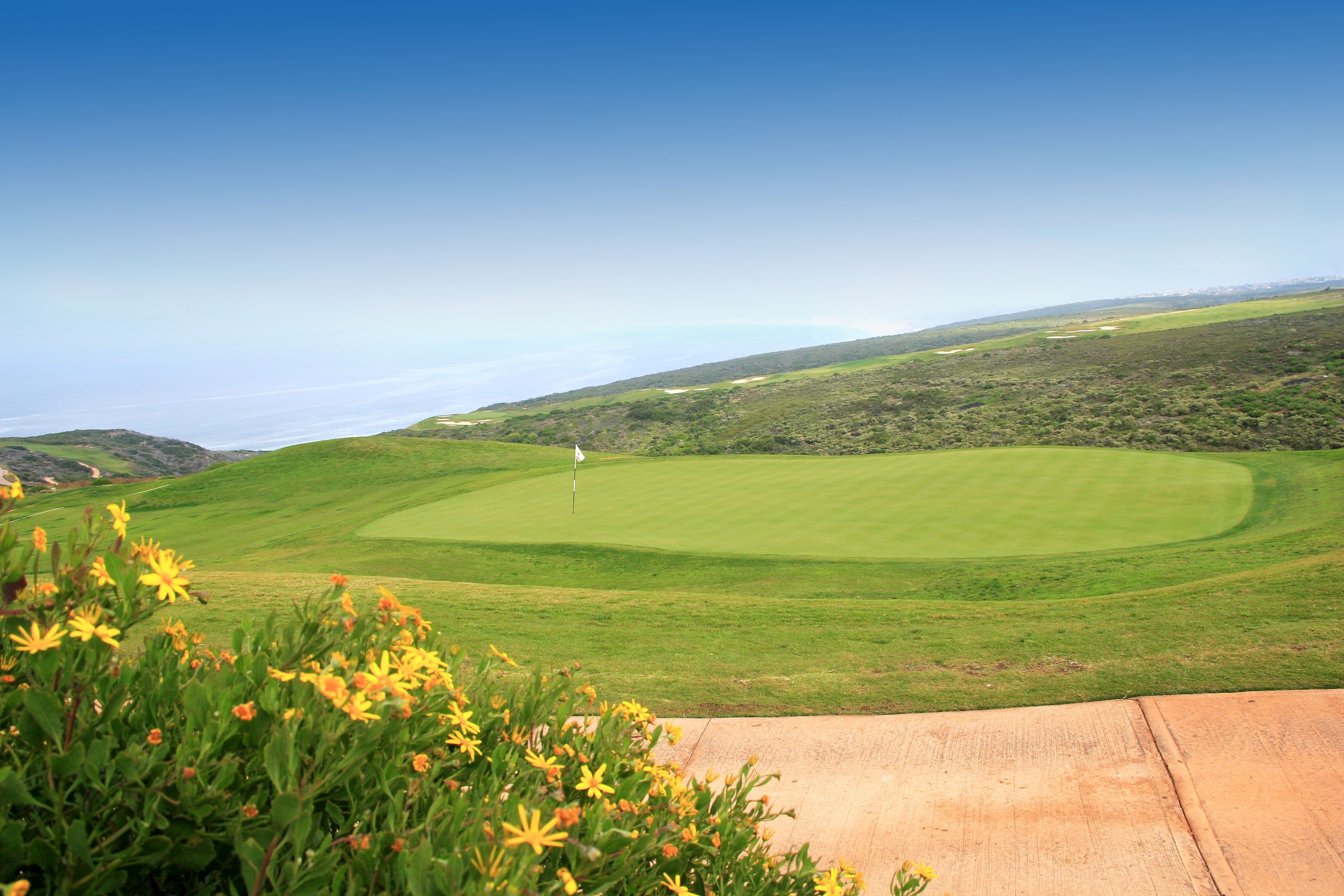 Un parcours de golf avec un drapeau sur le green, entouré de collines et sous un ciel dégagé.