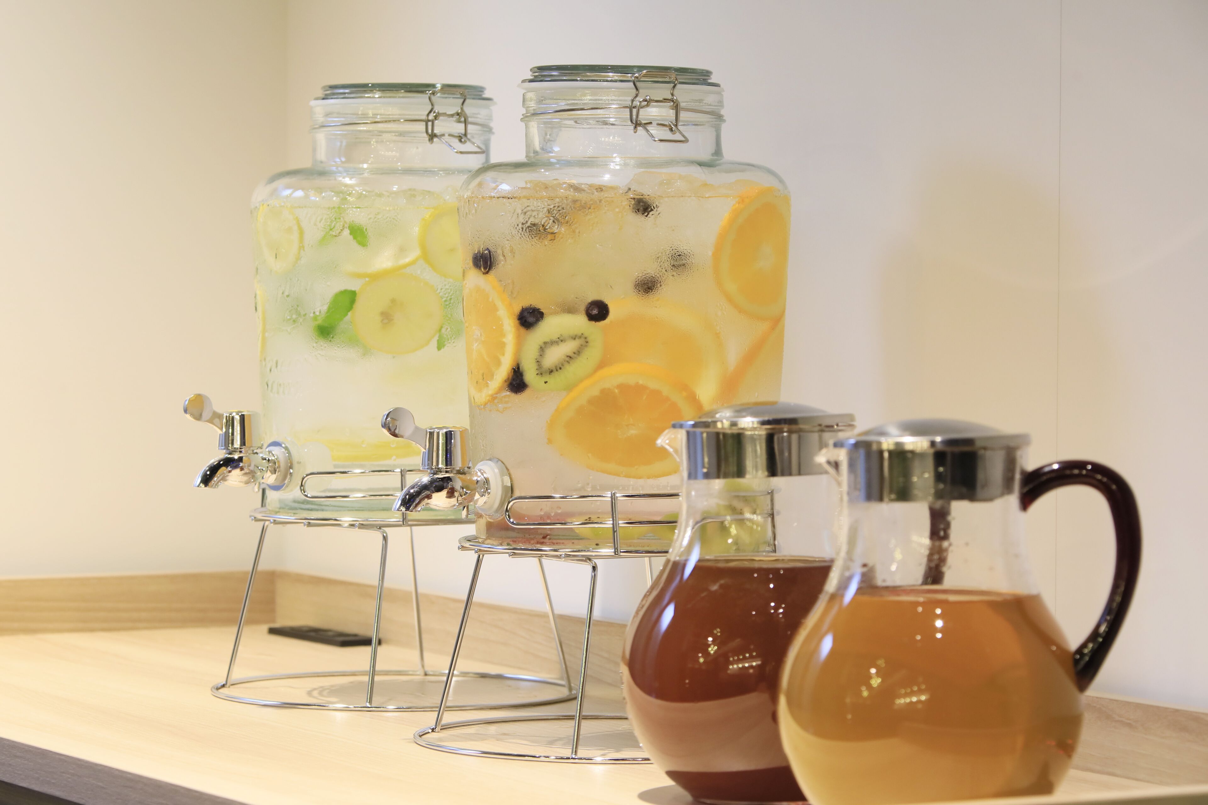 Three glass dispensers with fruit slices and a metal pitcher on a wooden surface.