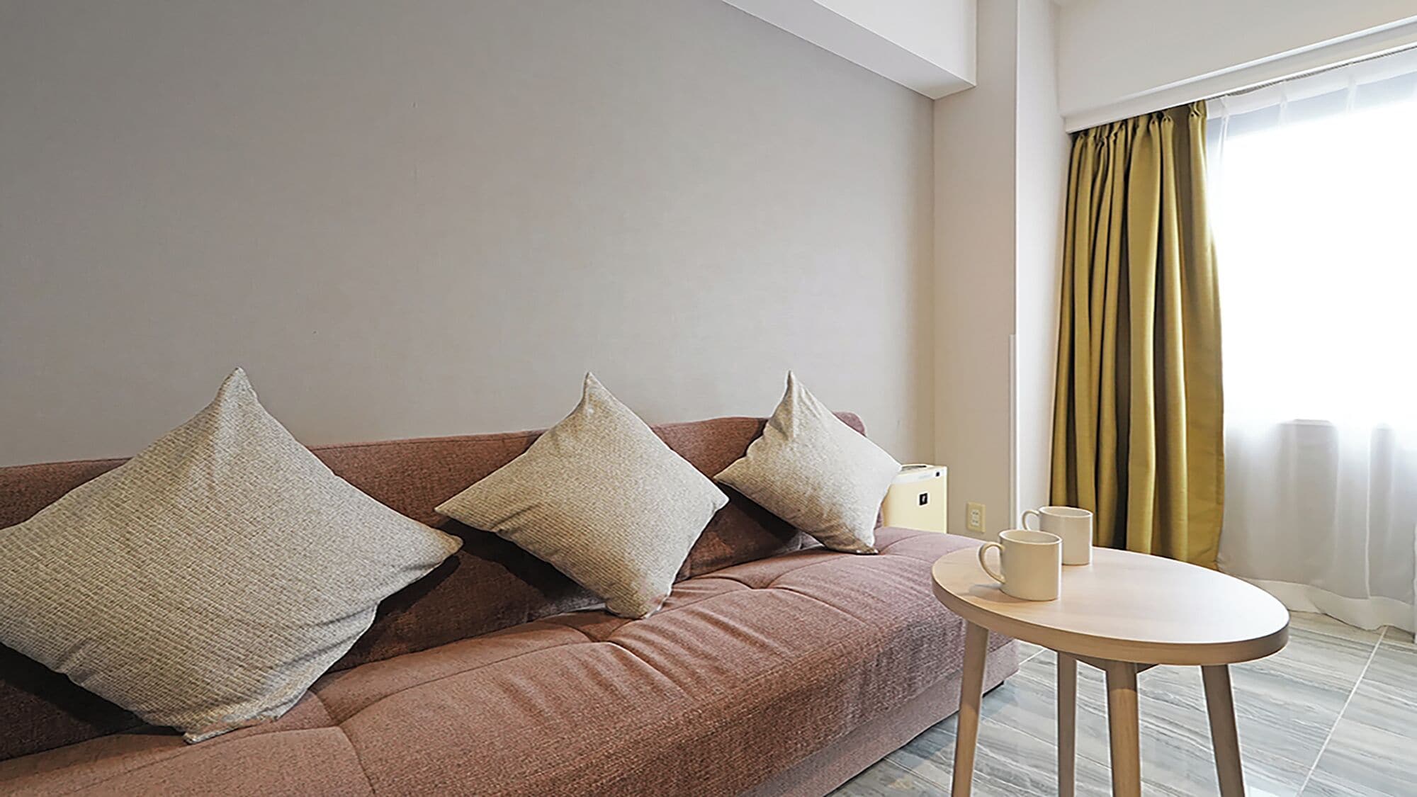 Ein modernes Wohnzimmer mit einem braunen Sofa, drei Kissen, einem kleinen Tisch mit zwei weißen Tassen und einem Fenster mit gelben Vorhängen.