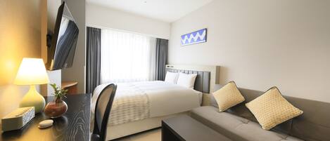 Habitación de hotel con cama, sofá, escritorio con lámpara, televisor en la pared y ventana con cortinas.