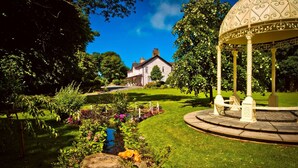 A gazebo with ornate metalwork, a well-maintained garden with various flowers, and a stone building in the background. - Plas Dinas Country House (Caernarfon)