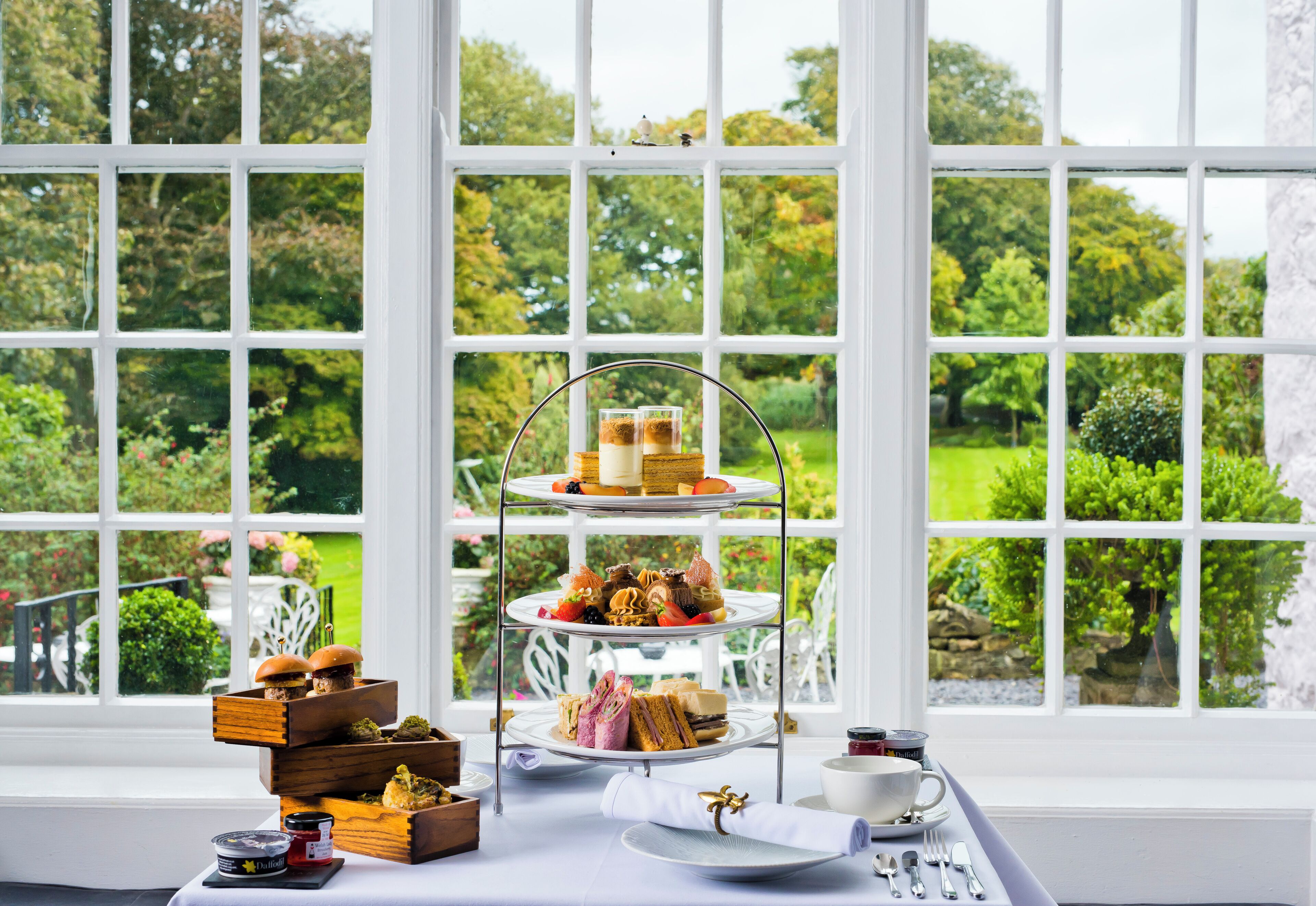 A three-tiered dessert stand with various pastries and sandwiches in front of a large window with a garden view.