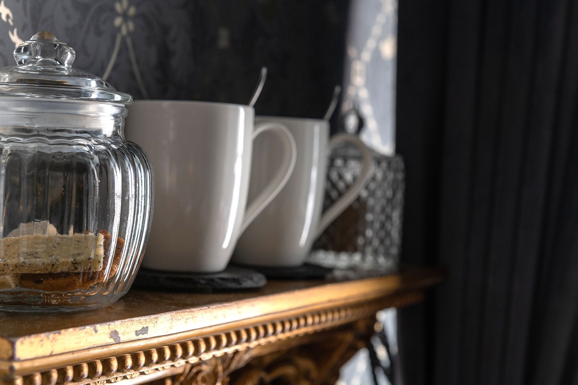 Ein Glas mit Deckel, eine weiße Tasse und ein verziertes Gefäß auf einer Holztischplatte.