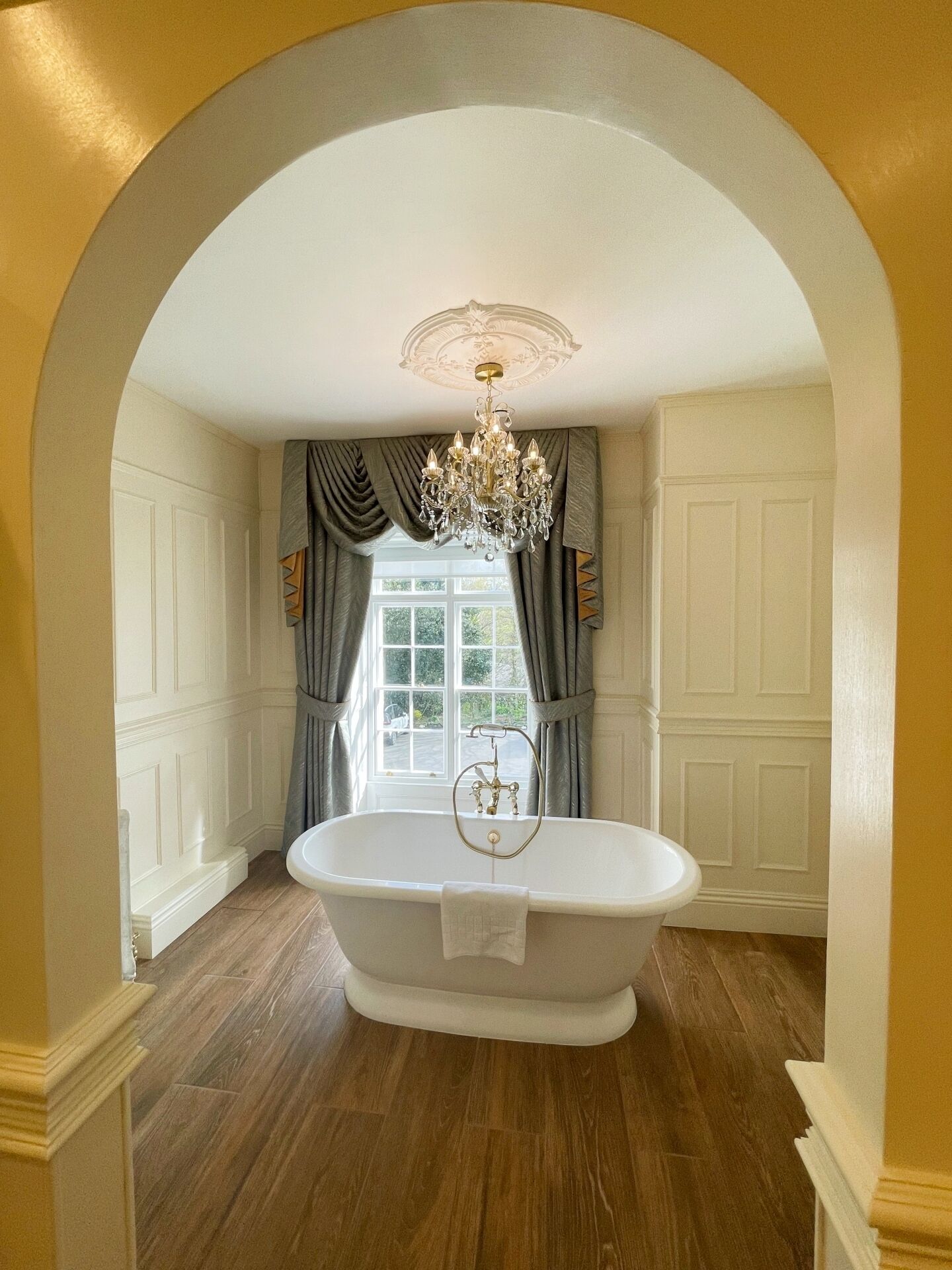 A bathroom with a freestanding bathtub, a chandelier, and a window with curtains.