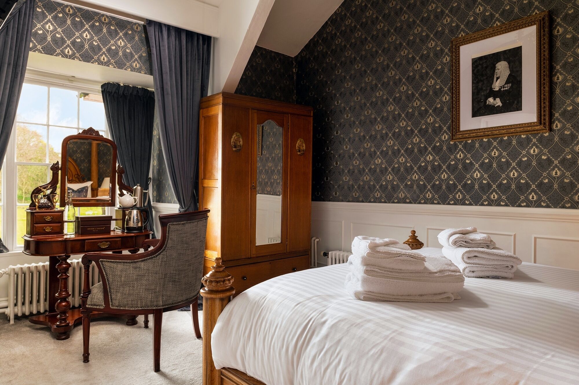 A bedroom with a large bed, a wooden wardrobe, a dressing table with a mirror, and a framed portrait on the wall.