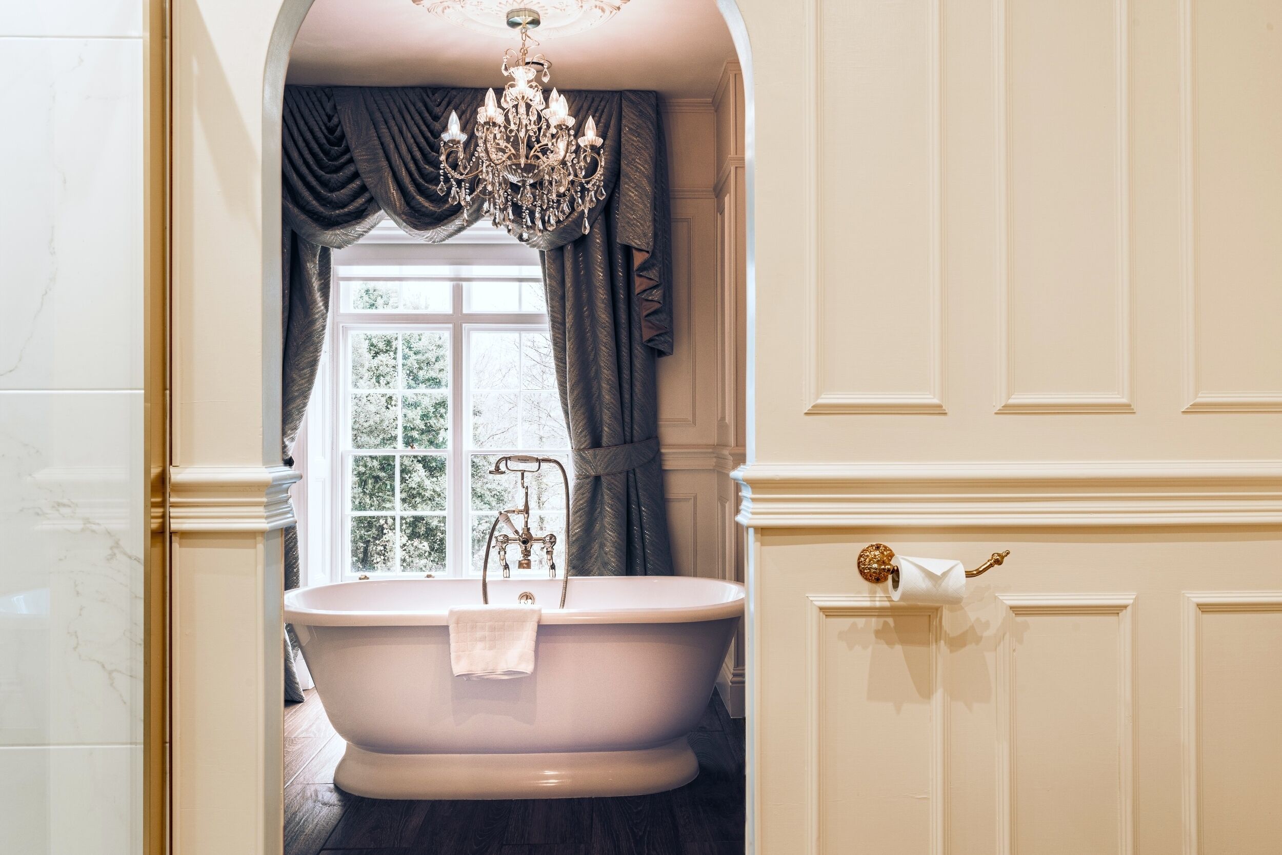 A bathroom with a freestanding bathtub, a chandelier, a window with curtains, and a toilet paper holder.