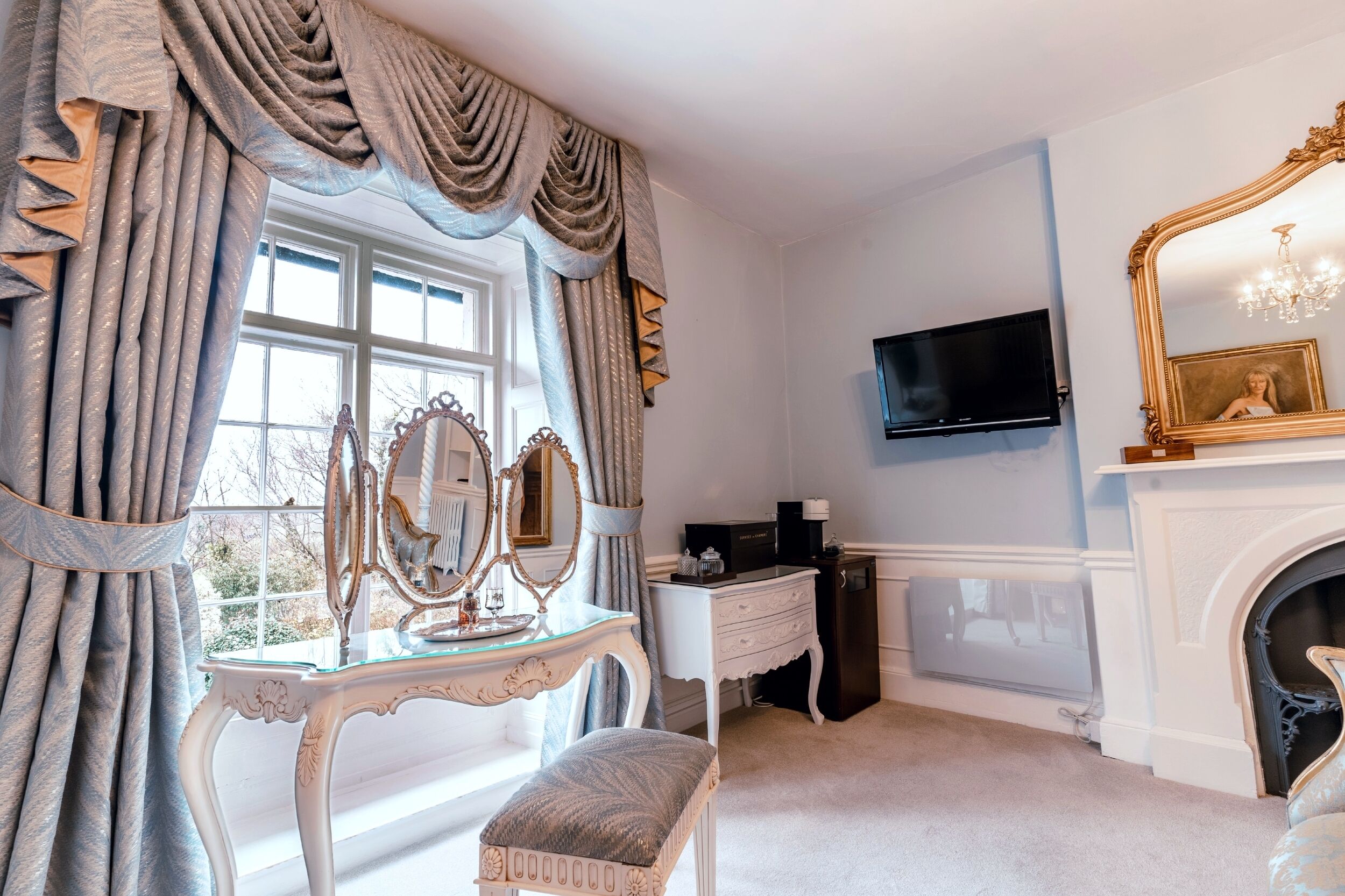 A room with a large window, a vanity with a mirror, a television mounted on the wall, and a fireplace.