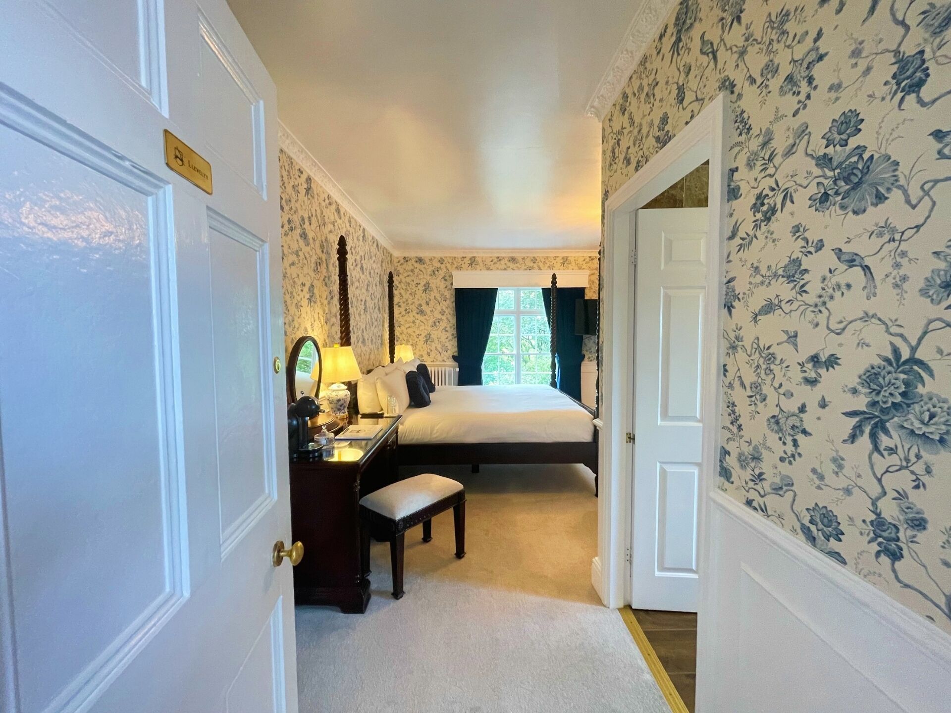 Quarto de hotel com cama, escrivaninha com luminária, cadeira e mesa pequena.