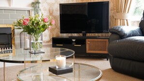 A living room with a glass coffee table, a black sofa, a flat-screen TV, a vase with flowers, and a fireplace. - The Sandpiper Guest House (Torquay)