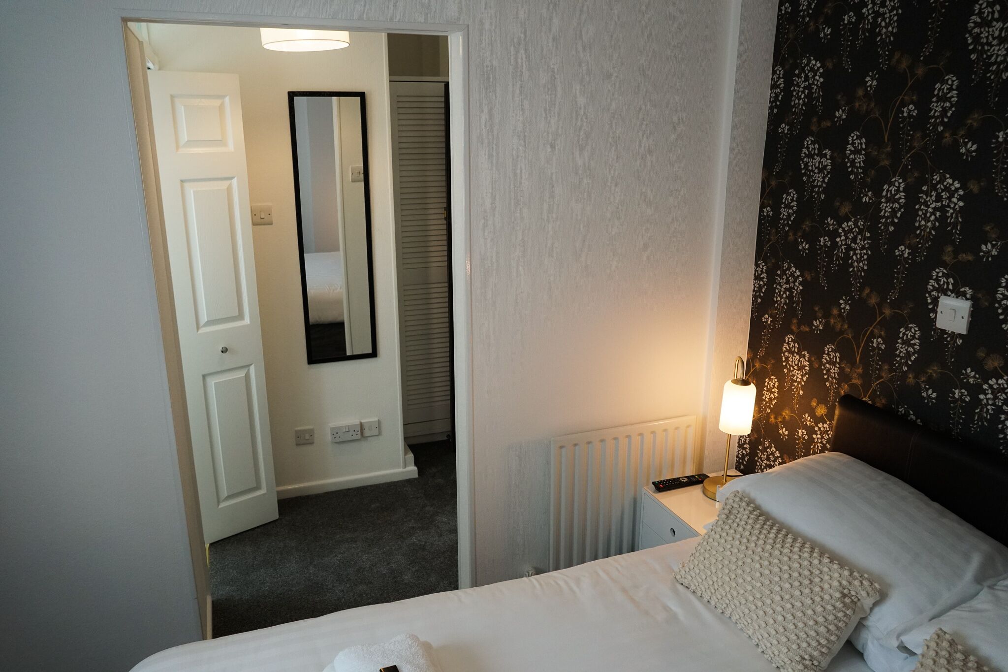 Une chambre d’hôtel avec un lit, une table de chevet, une lampe, un miroir et un radiateur.
