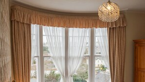 A neatly made bed with white linens and two pillows, a bedside lamp, and a chandelier. There are two chairs and a wooden cabinet in the room. - The Sandpiper Guest House (Torquay)